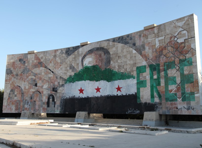 A large outdoor mural of a man's face is painted over with the Syrian revolution flag. Next to it, the English "Free" is painted in green. 