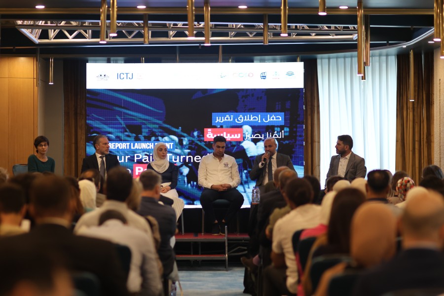 Six panelists sit in chairs on a stage facing a room full of people and the camera