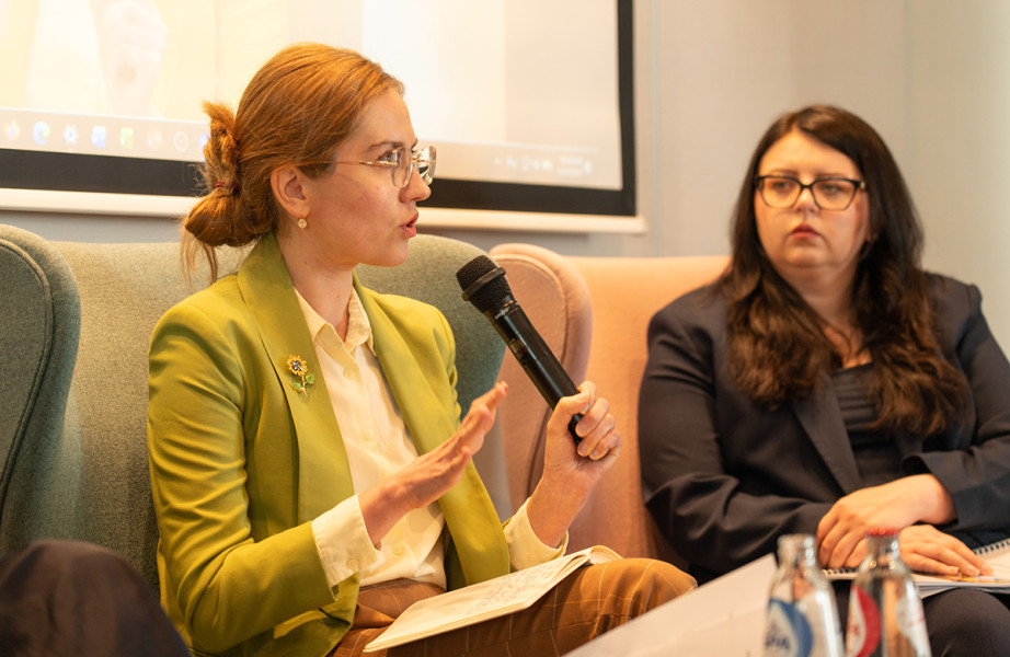 A women seated in chair (in profile position) speaks in a microphone.