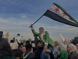 A large crowd of people in the street, many waving the Syrian flag
