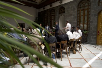 Some 20 people sit in chairs around a table located inside a courtyard.