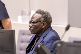 A man wearing a suit and glasses is seated in a chair, in profile position (side view), head looking straight ahead with a straight face.