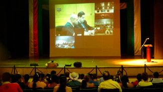  A seated audience watch formal proceedings projected on a screen mounted on a stage