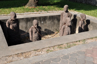 A sculpture featuring five human figures connected by chains and standing in a square hole in the ground