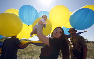A women holds up an infant in the air on sunny day outdoors, surrounded by yellow and blue balloons